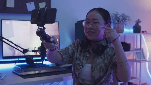 Waist up slowmo of Asian teenage streamer girl holding tripod with smartphone recording video blog of herself playing first person shooter game at home
