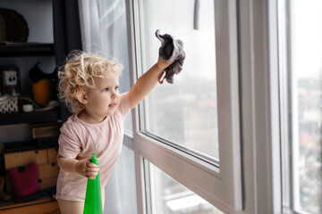 Housework. Cute curly child blonde girl helps to wash the windows in the apartment holding a towel...