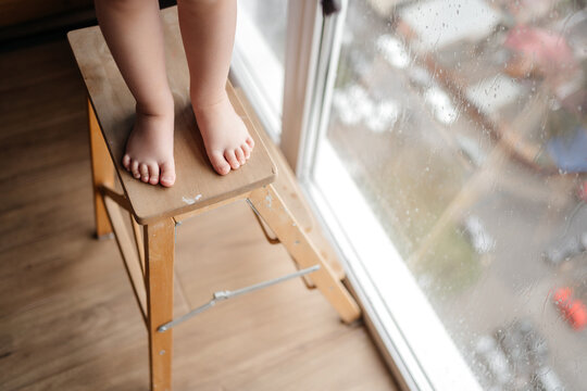 Legs of a small child on a ladder chair against a window with rainy weather. Attention Danger and children's interest in windows.