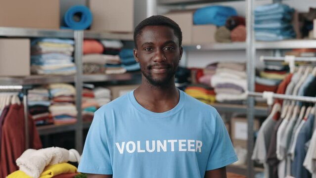Portrait view of the multiracial man in volunteer t shirt looking at the camera with smile. Shelves with belongings for donating at the background. Humanitarian aid concept