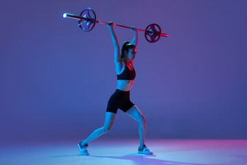 Portrait of muscled woman in sportswear training with a barbell isolated on purple background in neon light. Sport, weightlifting concept