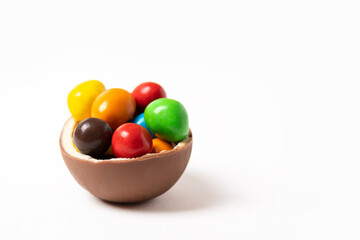 Cracked chocolate easter egg with colorful small round candies on white background, copy space, side view. Chocolate treat for kids. Easter concept.