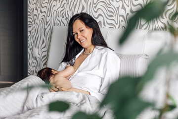 Portrait of smiling mother breastfeeding her infant daughter in bedroom, blurred foreground. Baby...