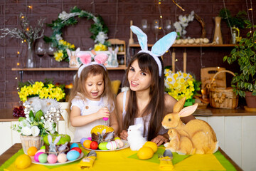 Easter, mom and daughter with Easter eggs and cake at the festive table are smiling and preparing for Easter at home in the kitchen in the ears of a hare.