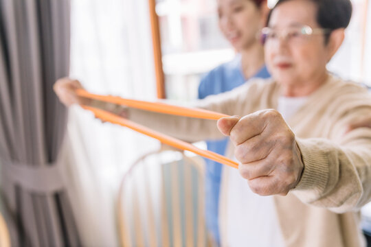 Close-up hand of a mature senior Asian woman using a resistance band with a nurse caregiver wearing scrubs, exercise for the senior patient in physiotherapy treatment. Home health care concept.