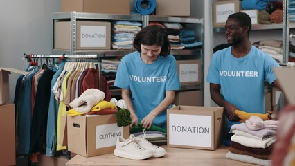 Brunette woman and multiracial man standing at the warehouse and sorting and iterating clothes for donations. Belongings at the shelves at the background. Humanitarian aid concept