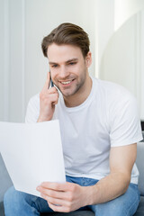 smiling man in white t-shirt looking at paper while talking on cellphone.