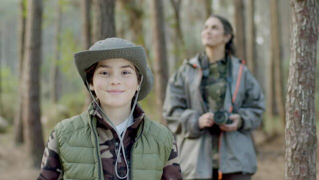 Happy mother and son exploring nature in forest on autumn day. Smiling boy with book wearing gray hat and green vest while woman standing and holding camera in background. Nature, leisure concept