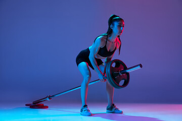 Studio shot of female athlete, sportive woman posing with barbell isolated on purple background in...