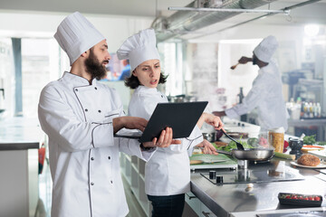 Head chef with modern laptop showing to sous chef recipe for gourmet dish required for gastronomic...