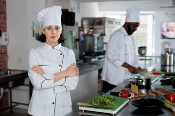 Portrait of confident restaurant kitchen worker standing with arms crossed while smiling at camera. Gastronomy expert wearing cooking uniform while preparing fresh herbs for dinner service.