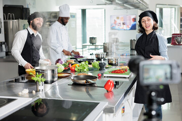 Fototapeta na wymiar Happy smiling chefs shooting culinary video course in restaurant kitchen while preparing delicious dish. Multiethnic cooks recording food preparation process for television cooking show.