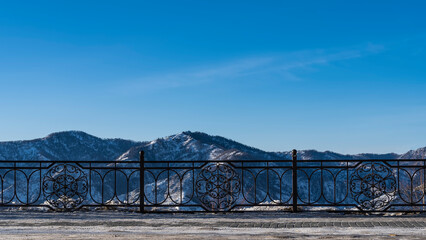 A metal fence is installed along the road. Through the openwork bars, snow-capped mountains can be seen against the blue sky. Copy space. Altai