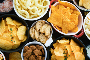 Unhealthy food or snacks. All classic potato snacks with peanuts, popcorn and onion rings and salted pretzels in bowl plates on old wooden background. Unhealthy food for figure, heart, skin, teeth.