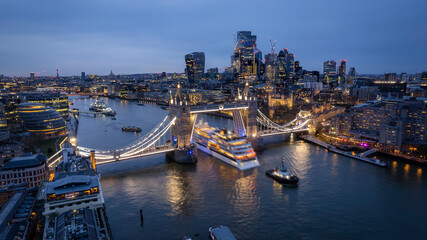 Fototapeta na wymiar Aerial view of the skyline of London with a motion blurred cruise ship passing under the lifted Tower Bridge during dusk, England