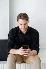 stylish man in black shirt sitting at home and messaging on smartphone.