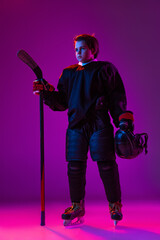 Full-length portrait of little boy, child, hockey player in special uniform posing with stick isolated over purple background in neon light