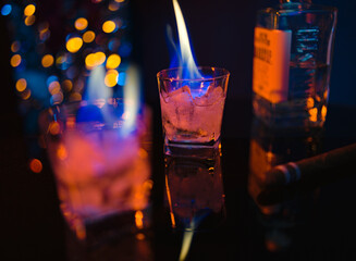 Fire and ice on the bar. Two glasses of strong alcohol with ice, a cigar, a bottle and bar lights in a blurry background. Flames in glasses with a strong drink with ice.
