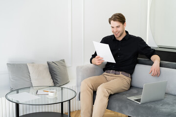 cheerful man reading document while sitting on couch near laptop and coffee table.