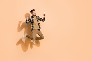 Full length body size view of handsome trendy cheery guy jumping using device having fun isolated on beige pastel color background