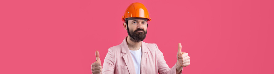 Portrait of young construction engineer wear orange hard hat, in a pink jacket standing on red studio background. The man points with his hand.