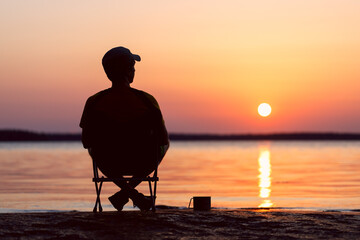 Happy man is sitting on a camping chair with a mug of tea on the shore of a beautiful evening...