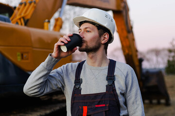 Builder in helmet drinking coffee from paper cup on coffee break. Excavator on the background.