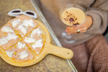 Woman holding wooden board with small pizza and glass of shakerato drink at outdoor restaurant, close-up, cropped view focused on pizza. Concept of italian cuisine while travel