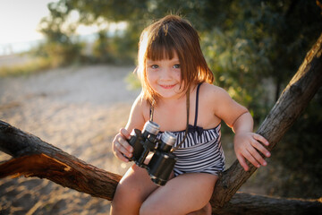 Cute european child girl in a bathing suit with binoculars on a tree. Summer holidays and adventures. Jungle games on the sea coast.