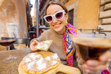 Woman having fun while eating pizza and drinking cocktail at restaurant on a street in Rome. Concept of Italian gastronomy and travel. Stylish woman with sunglasses and colorful hair shawl
