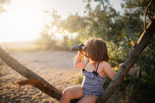 Cute european child girl in a bathing suit with binoculars on a tree. Summer holidays and adventures. Jungle games on the sea coast. - Powered by Adobe
