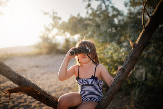 Cute european child girl in a bathing suit with binoculars on a tree. Summer holidays and adventures. Jungle games on the sea coast. - Powered by Adobe