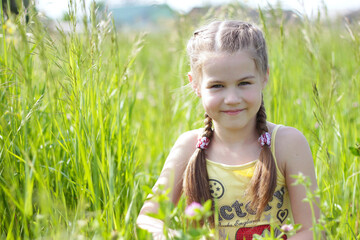 A little girl with pigtails sits on green grass in a summer park on a sunny day