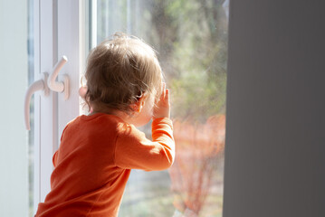baby girl is looking through window glass at sunny day, touching the glass and having a fun with...