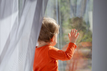 baby girl standing in front of the window glass at sunny day, touching the glass and having a fun with the sunbeams, natural dirty glass and curtain - obrazy, fototapety, plakaty