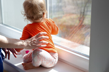 baby girl sitting on windowsill in front of the window glass at sunny day, mother holds child, natural dirty glass - obrazy, fototapety, plakaty