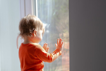 baby girl standing in front of the window glass at sunny day, touching the glass and having a fun with the sunbeams, natural dirty glass
 - obrazy, fototapety, plakaty