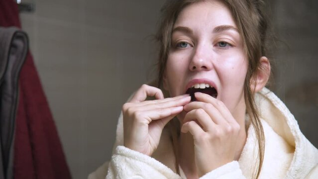 Attractive young Caucasian woman in a bathrobe standing in the bathroom installs a polymer aligner on her lower jaw. Oral care and bite restoration and teeth alignment
