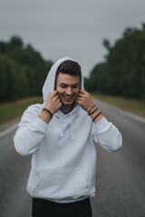 A young handsome guy with a small beard in a white jacket walks along the road puts on a hood and smiles