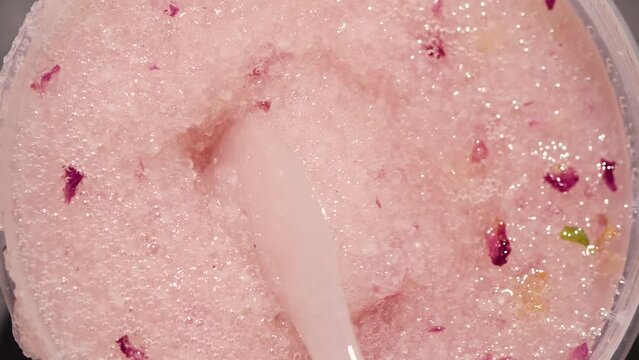 Glass jar with wet viscous body scrub made of sea salt and rose petals stirred with a white spatula. Close-up. High quality 4k footage