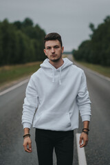 A young handsome guy with a small beard in a white jacket walks along the road and looks at the camera