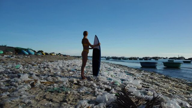 Caucasian young athletic female surfer stand in polluted beach with plastic waste trash garbage, Mother Earth ocean pollution save the planet concept