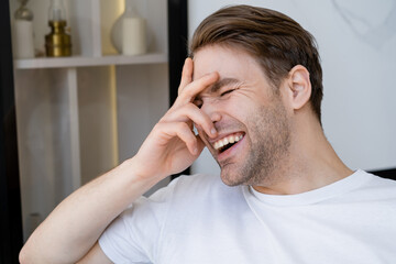 excited man in white t-shirt obscuring face with hand while laughing at home.
