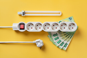 Power strip on 100 euro banknotes, electric plug and fluorescent light bulb over yellow background....