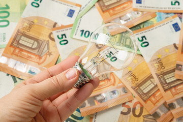 Woman hand holding light bulb over euro banknotes background. High electricity costs due to power...