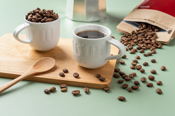 Cup of freshly brewed coffee, spoon and scattered coffee beans on a pastel green background. Whole organic coffee beans spilled out of a brown paper bag. Making tasty strong coffee concept.