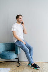 full length view of smiling man in jeans talking on smartphone near armchair and grey wall.