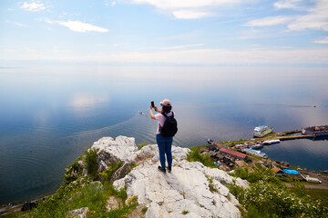 Fototapeta premium A female traveler stands on a rock near the port of Baikal and photographs Lake Baikal on a summer day. The concept of freedom, hiking.