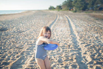 Cute european child girl in a swimsuit runs on the sand and plays frisbee on the beach. Summer holidays and adventures. Active games on the sea coast.