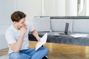 thoughtful man with pencil and paper sitting with crossed legs near laptop with blank screen.